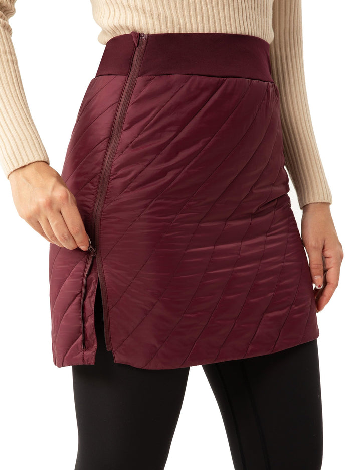 A close-up of a model wearing a quilted, wrap-style mini skirt in burgundy. The skirt features a diagonal quilted pattern, a front zipper, and a side slit that the model is pulling open.