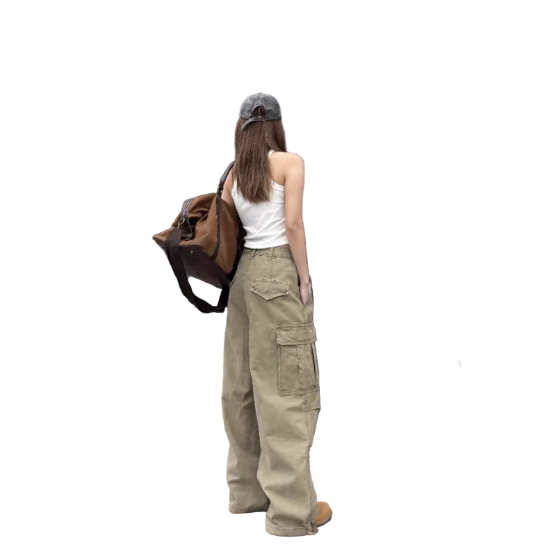 A woman stands with her back to the camera wearing wide-leg khaki cargo pants, a white tank top, and a gray cap, carrying a brown shoulder bag in front of a storefront.
