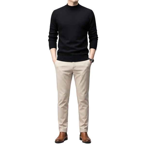 A man wearing a black long-sleeve mock neck knit sweater and light beige trousers, standing against a dark gray studio background.