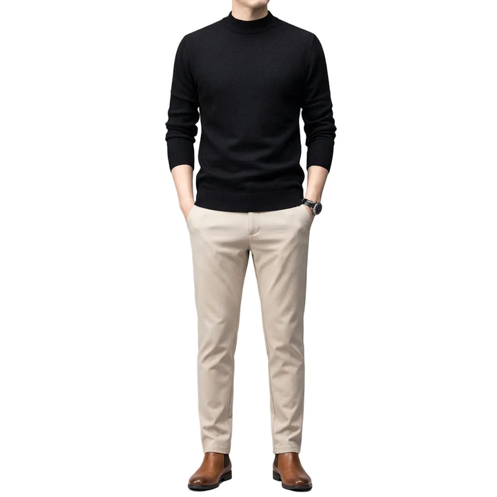 A man wearing a black long-sleeve mock neck knit sweater and light beige trousers, standing against a dark gray studio background.