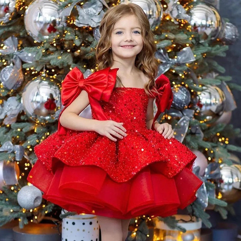 A young girl with wavy light brown hair stands in front of a decorated Christmas tree wearing a bright red sequined party dress with large bow details on the shoulders.
