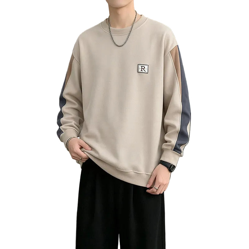 A man wearing a beige crewneck sweatshirt with dark gray and brown color-blocked sleeves, featuring a small embroidered letter 'R' on the chest, paired with black pants in a modern living room setting.