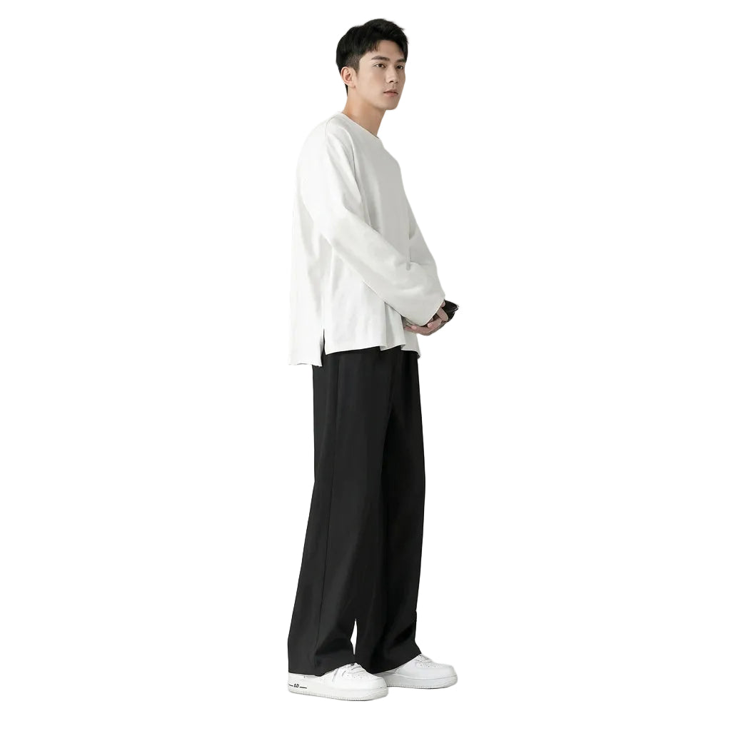 A model from the waist down wearing black wide-leg trousers, a white long-sleeve shirt, and white sneakers, standing against a solid muted olive-gray background.