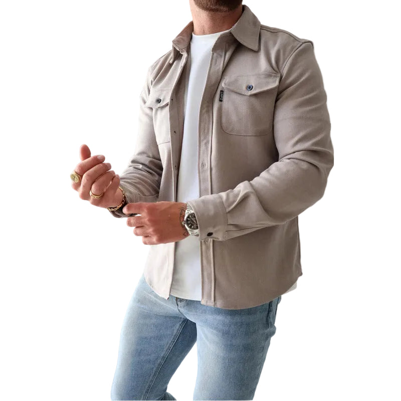 A man wearing a light beige long-sleeve button-up shirt jacket over a white t-shirt and blue jeans, shown from the chin down in a studio setting.