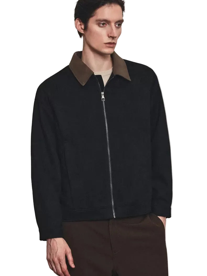 A man wearing a black zip-up jacket with a contrasting brown collar, layered over a light beige shirt and paired with brown trousers, standing against a textured light gray wall.