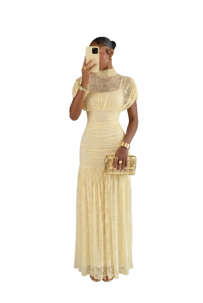 A woman takes a mirror selfie wearing a cream lace maxi dress with a high mock neck, short puff sleeves, and a fitted ruched bodice. She is holding a gold clutch in front of a dark wood door.