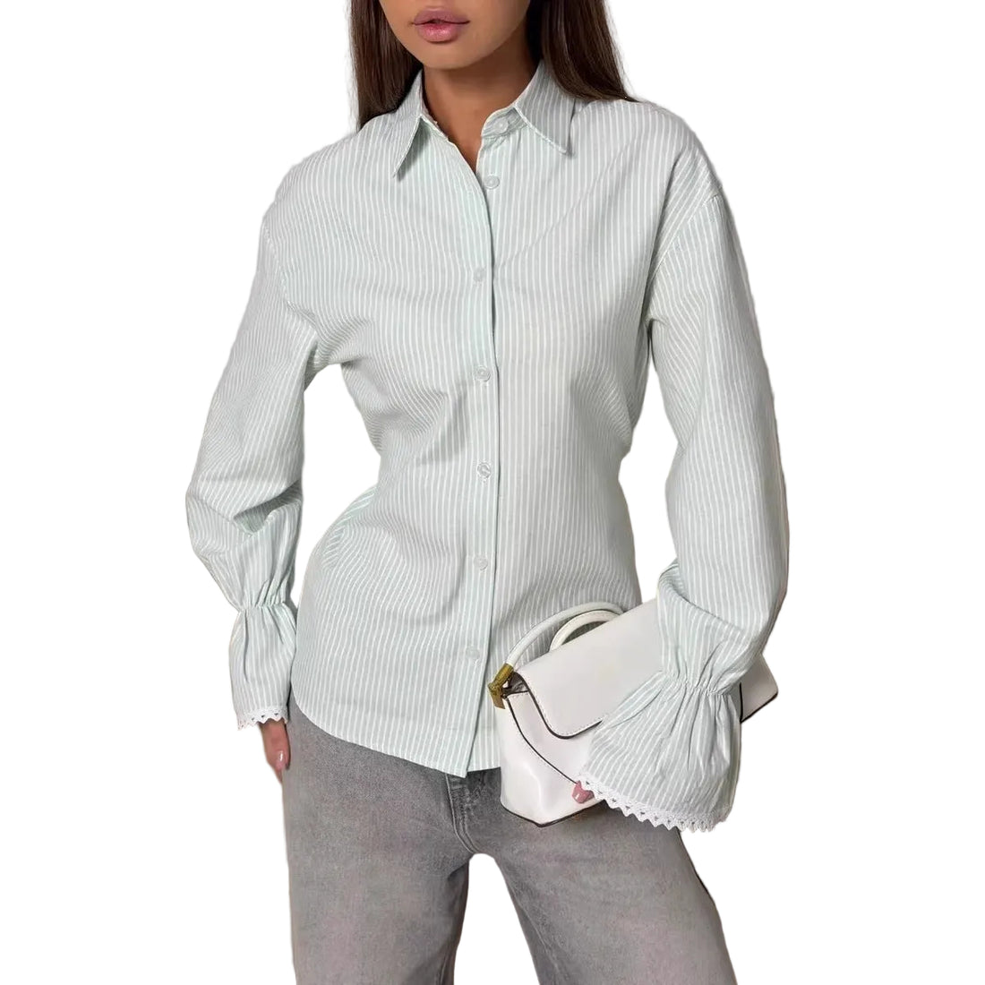 A woman wearing a light sage green and white vertically striped long-sleeve button-up shirt with gathered cuffs, paired with gray jeans and holding a white handbag, posed in front of a beige sofa.