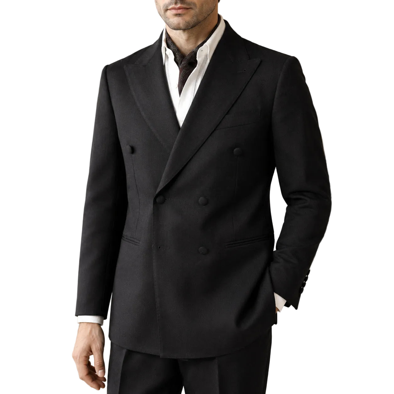 A man wearing a black double-breasted suit jacket with peak lapels, paired with a white dress shirt and dark tie, standing in an elegant indoor setting with a lamp in the background.