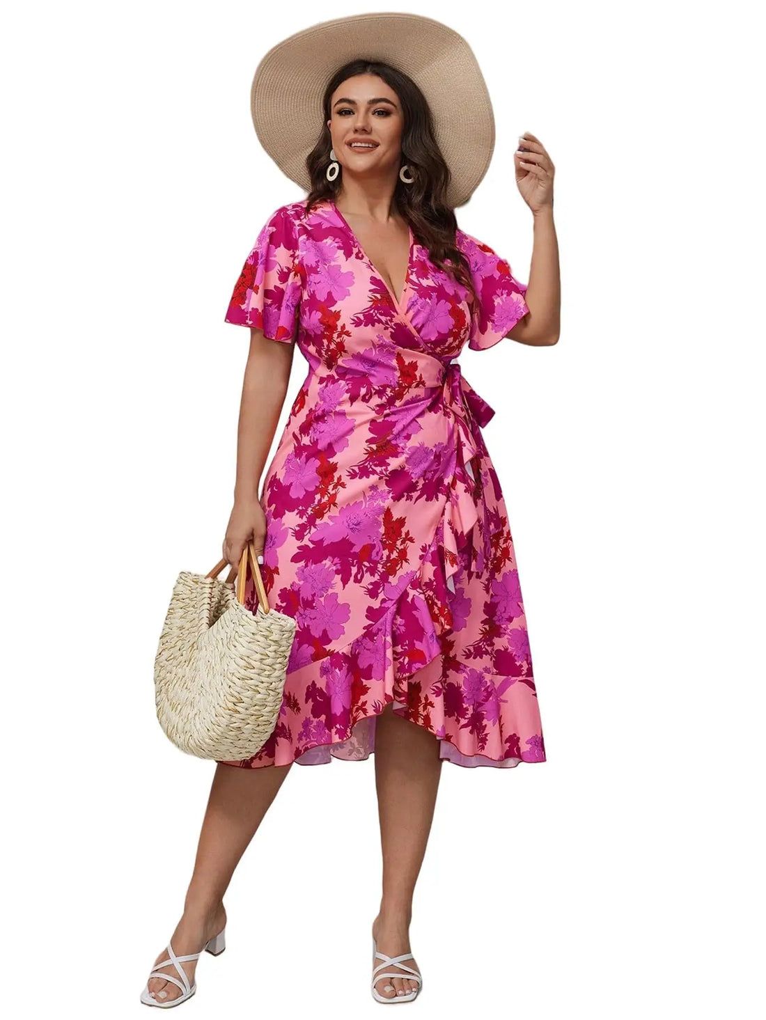 A woman modeling a pink and magenta floral print wrap dress with short flutter sleeves, a V-neckline, and a self-tie waist, styled with a wide-brimmed beige hat, a woven straw tote bag, and white heeled sandals against a plain white background.