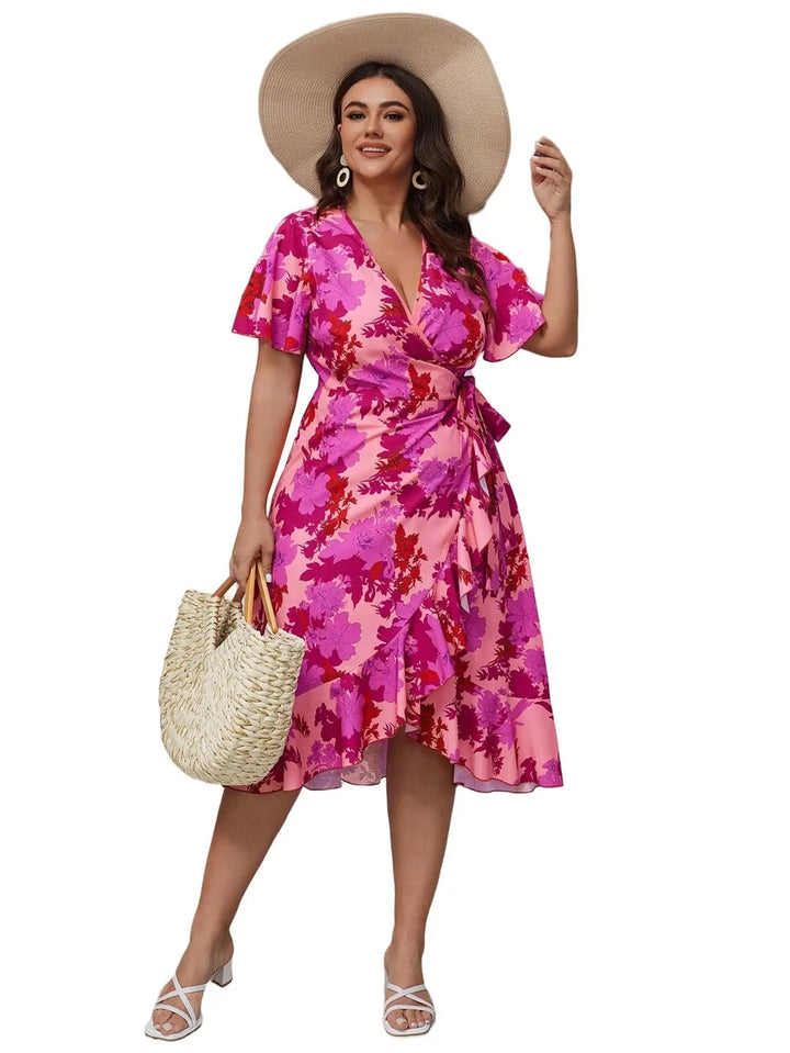 A woman modeling a pink and magenta floral print wrap dress with short flutter sleeves, a V-neckline, and a self-tie waist, styled with a wide-brimmed beige hat, a woven straw tote bag, and white heeled sandals against a plain white background.