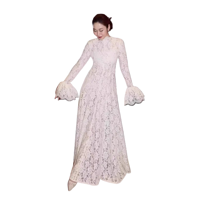 A woman wearing a long-sleeved, high-neck lace maxi dress with dramatic flared bell sleeves, standing in an indoor setting with a dark cabinet and a bench in the background.