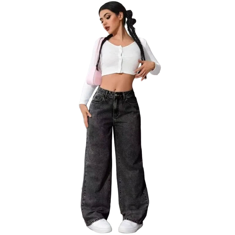 A woman stands against a gray concrete wall wearing a white cropped cardigan with light pink sleeves, dark gray wide-leg jeans, and white sneakers.