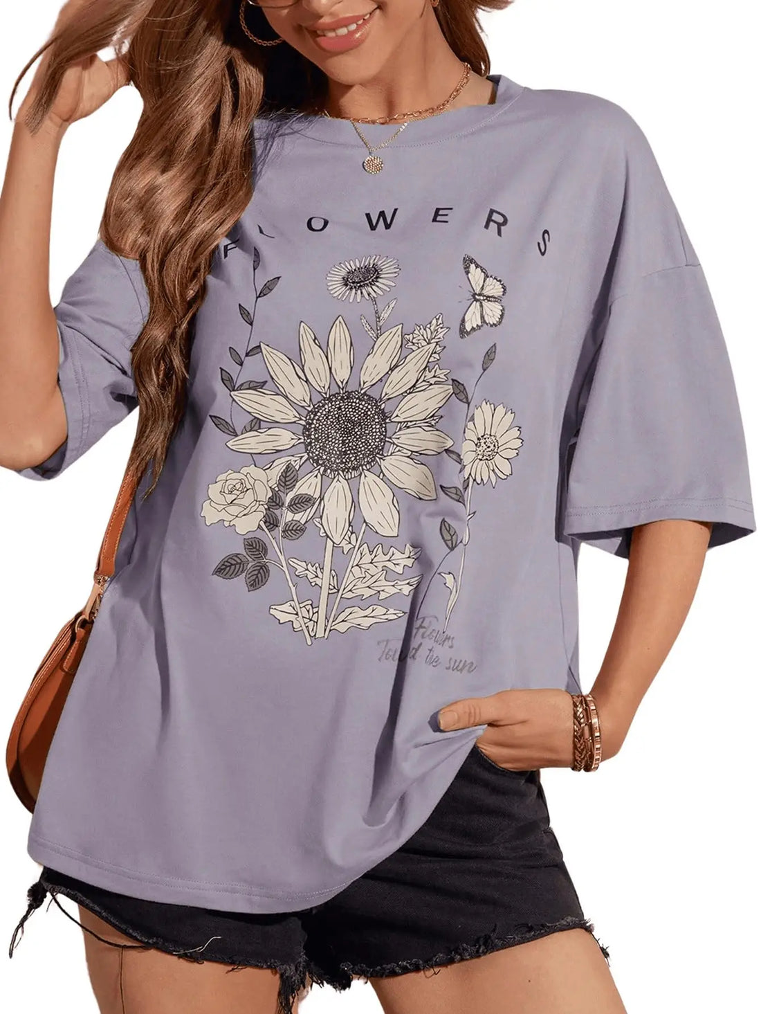 A woman wearing an oversized lavender-gray graphic T-shirt with a large sunflower, smaller flowers, leaves, and a butterfly print, paired with black denim shorts. The shirt has the word 'FLOWERS' printed across the chest.