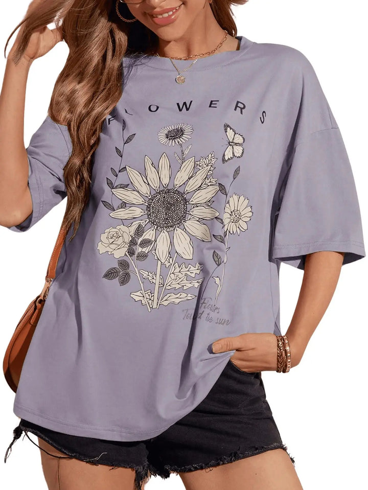 A woman wearing an oversized lavender-gray graphic T-shirt with a large sunflower, smaller flowers, leaves, and a butterfly print, paired with black denim shorts. The shirt has the word 'FLOWERS' printed across the chest.