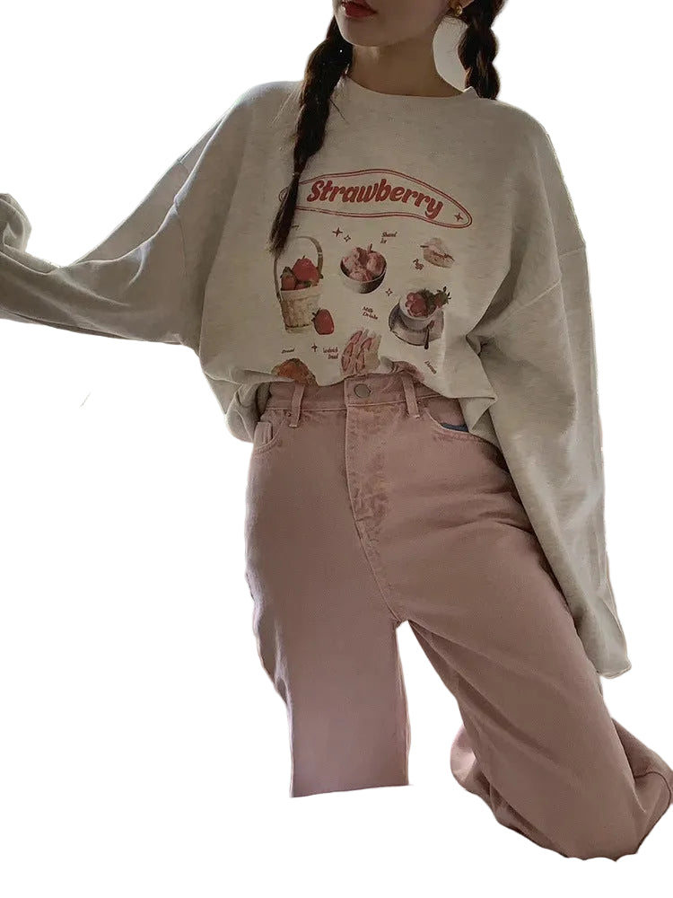 A person wearing a light beige oversized sweatshirt with a strawberry graphic print and pink high-waisted jeans, standing in front of a window.